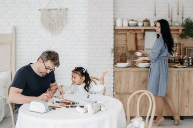 Father and daughter bond over crafts while mother prepares food in a cozy home setting.